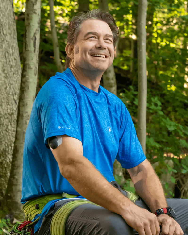 Ein lächelnder Mann, der ein blaues Shirt und ein Eversense Glukosermessgerät trägt, sitzt in einem sonnendurchfluteten Wald.