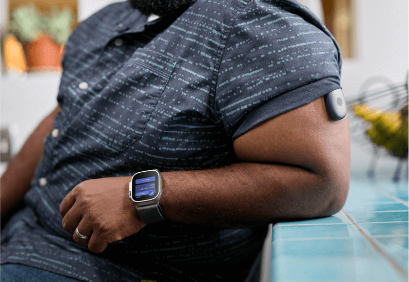Eversense Benutzer mit Smartwatch und Arm-CGM-Sensor in einer Küche, an den Tresen gelehnt.