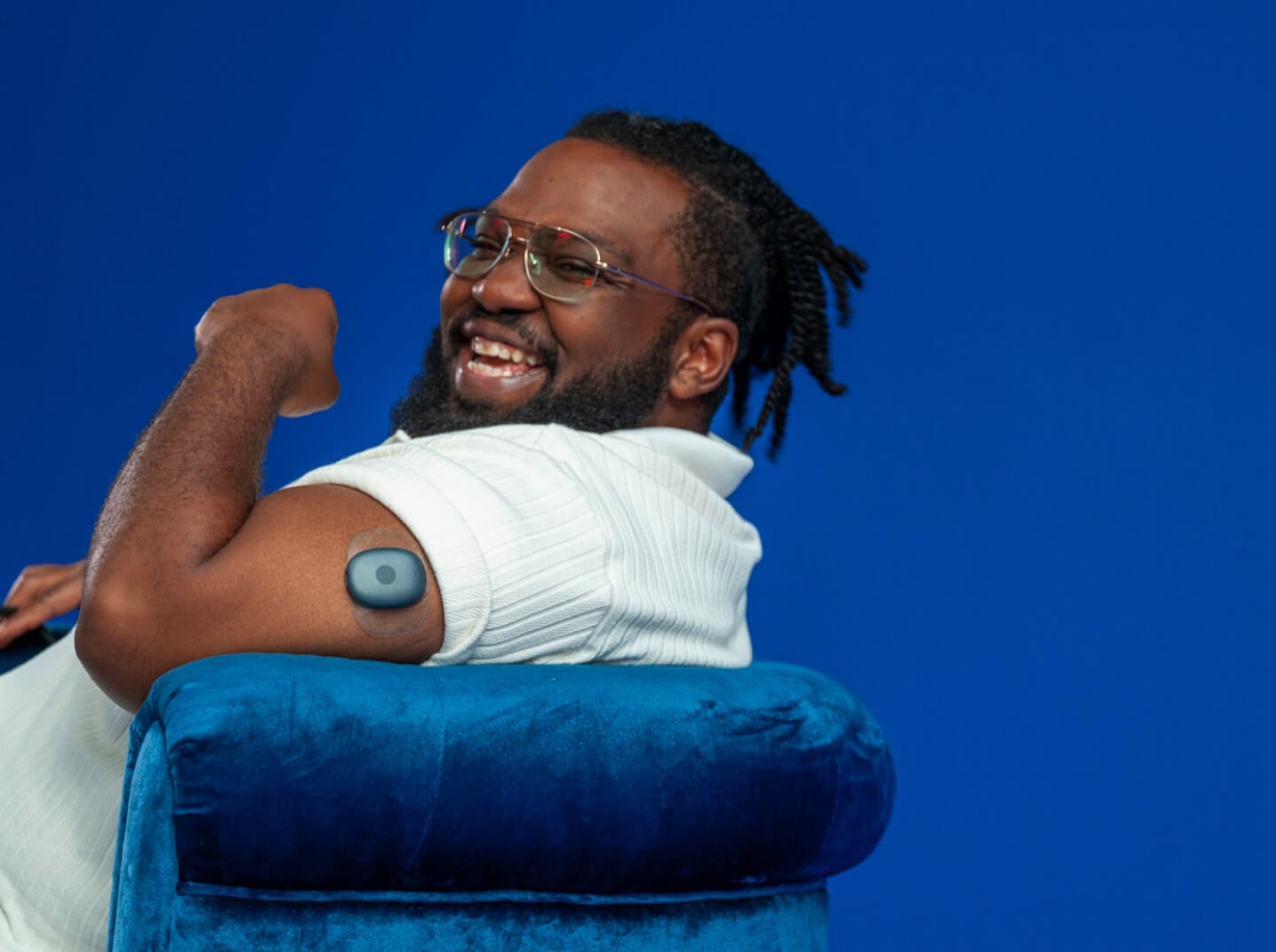 Lächelnder Eversense Benutzer mit gebeugtem Arm, an dem ein Eversense Transmitter angebracht ist, auf einem blauen Stuhl sitzend.