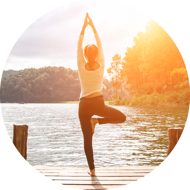 Eversense Benutzerin beim Üben der Yoga-Baumstellung auf einem hölzernen Steg, umgeben von einem ruhigen See und bewaldeten Hügeln, mit Sonnenlicht.