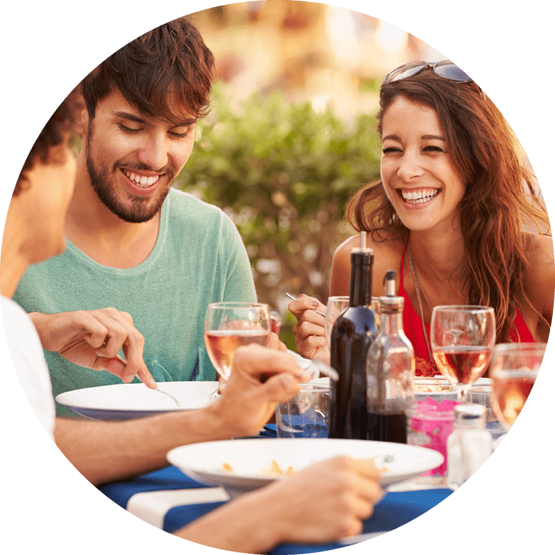 Drei Personen lächeln und essen gemeinsam an einem Tisch im Freien, umgeben von Getränken, Flaschen und verschwommenem Grün.