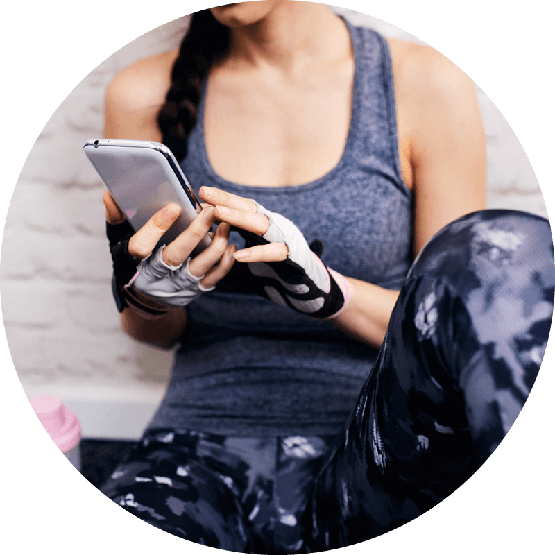 Eversense Benutzerin mit Smartphone in der Hand, in Trainingskleidung und mit fingerlosen Handschuhen, sitzt drinnen vor einer weißen Backsteinwand.