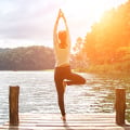 Eversense Benutzerin beim Üben der Yoga-Baumstellung auf einem hölzernen Steg, umgeben von einem ruhigen See und bewaldeten Hügeln, mit Sonnenlicht.