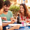 Drei Personen lächeln und essen gemeinsam an einem Tisch im Freien, umgeben von Getränken, Flaschen und verschwommenem Grün.