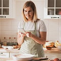 Eine Eversense CGM Benutzerin in einer Schürze benutzt ein Smartphone in einer hellen Küche, in der Backzutaten auf der Theke liegen.