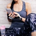 Eversense Benutzerin mit Smartphone in der Hand, in Trainingskleidung und mit fingerlosen Handschuhen, sitzt drinnen vor einer weißen Backsteinwand.